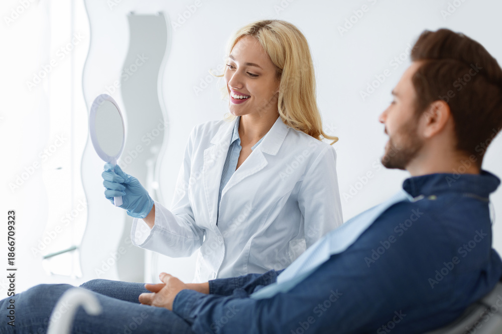 Obraz premium Pretty young blonde woman dentist holding mirror for male patient sitting in dental chair, stomatologist showing results of treatment, cheerful barded man looking at mirror and smiling, copy space