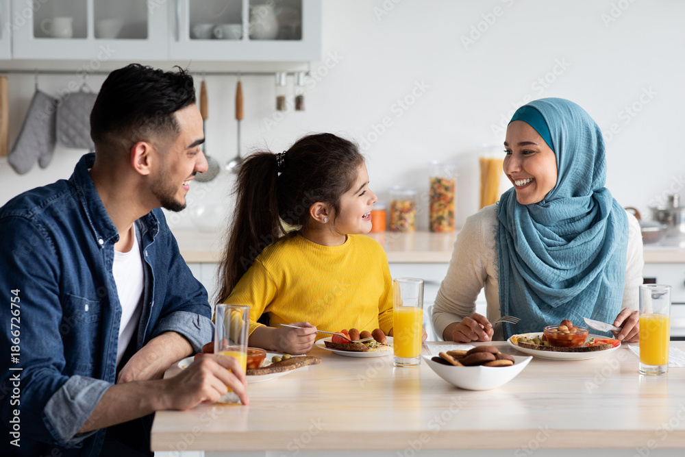 Fototapeta premium Domestic Breakfast. Happy Middle Eastern Family Of Three Eating Together In Kitchen, Cheerful Young Arab Parents And Little Daughter Sitting At Table, Enjoying Tasty Food, Chatting And Laughing