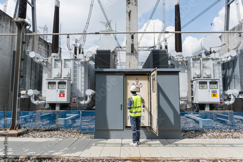 Bild auf Leinwand Male electrical engineer inspecting control cabinet of large power transformers at high voltage substation to monitor energy distribution grid and industrial utility infrastructure
