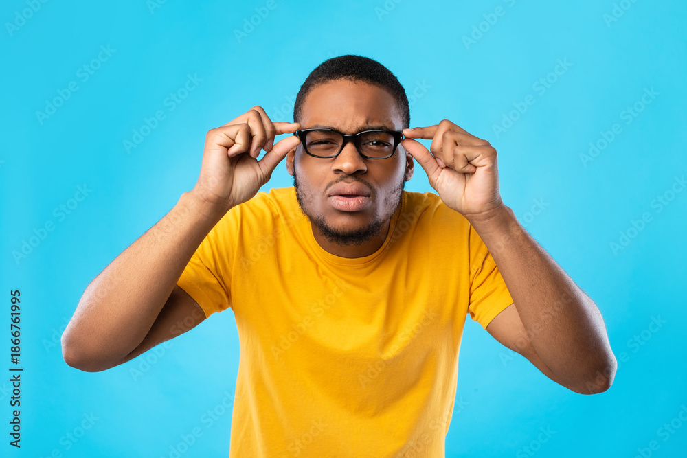 Fototapeta premium African American Guy With Poor Eyesight Wearing Eyeglasses And Squinting Eyes Looking At Camera Through Glasses Standing Over Blue Studio Background. Healthcare, Vision Problems