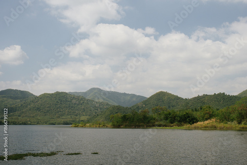 Beautiful outdoor view at Huai Tha Khoei Ban Kha Ratchaburi reservoir. The scene features a wooden photo spot, white ducks, and a calm lake with mountain views.
