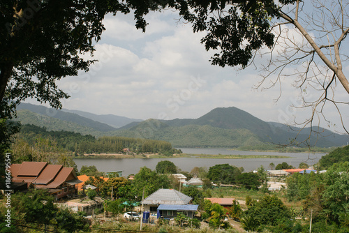 Beautiful outdoor view at Huai Tha Khoei Ban Kha Ratchaburi reservoir. The scene features a wooden photo spot, white ducks, and a calm lake with mountain views.