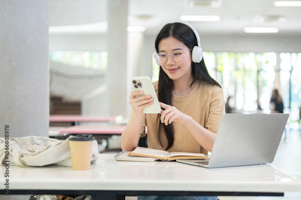 Obraz premium Glasses asian woman college student listening to music and looking at phone sit at table in building