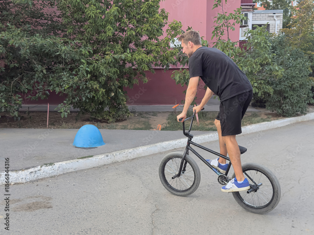Fototapeta premium A person rides a BMX bike on a street while trees surround the area and a blue object is nearby