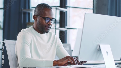 A man works intently at a sleek computer in a stylish office space. Natural light fills the room, enhancing the productive atmosphere. The scene reflects a contemporary business setting.