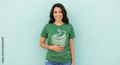 A smiling brunette woman wearing a green t-shirt with a digestive system graphic stands against a light blue wall in natural lighting. She is holding her abdomen area.