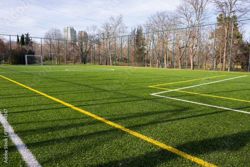 Foto an expansive green artificial turf soccer field marked with white and yellow lines, enclosed by a tall wire fence and surrounded by bare trees and distant city buildings