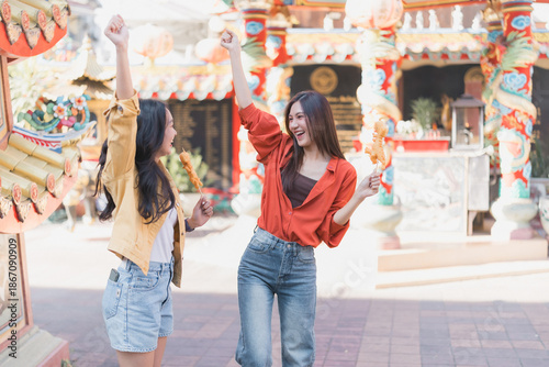 Tableau sur toile Two young Asian women joyfully raising arms and holding snacks on sticks, explor