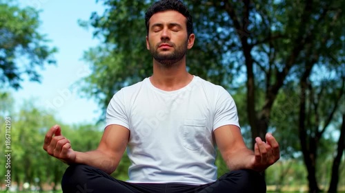 A man sits in a meditative pose outdoors, eyes closed, amidst trees
