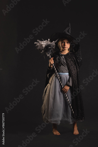 Child dressed as witch holding decorative staff in dark studio, wearing hat, cape and tulle skirt - costume for Halloween or carnival