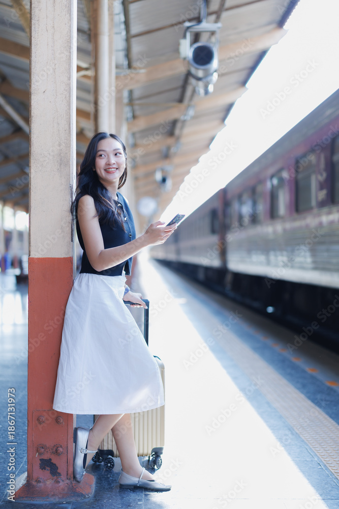 Fototapeta premium Asian girl standing next to her luggage on a train station platform, smiling and checking her smartphone while waiting for the train to arrive, anticipating her journey