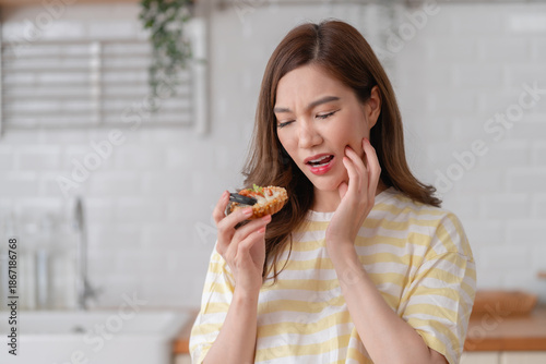 Foto Young asian woman wincing in pain and touching her cheek while holding a sweet tart, suffering from tooth sensitivity or dental problems in a bright kitchen