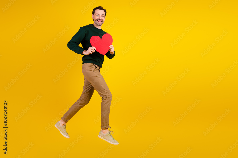 Fototapeta premium Happy young man in casual sweater jumps holding a red heart against a bright yellow background for love and lifestyle promo