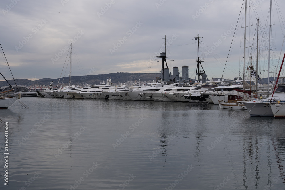 Fototapeta premium Modern marina in Athens, Greece with luxury yachts lined along the pier. Calm harbor water, sleek hulls and cloudy sky form a clean maritime travel scene.