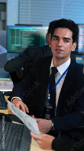 Project manager discussing data reports with a colleague in the office. Software code on monitors highlights a collaborative corporate IT and analytics environment of developer workspace.