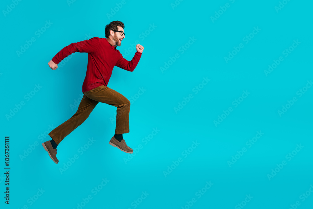 Fototapeta premium Energetic young man in red sweater and brown pants jumps in air blue background for dynamic stock photography about fitness style lifestyle and advertising appeal