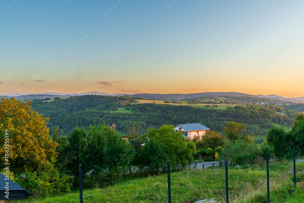 Obraz premium Wide countryside panorama at sunset with layered green hills, scattered houses, and a soft gradient sky, creating a peaceful scenic background for travel, environment, and rural lifestyle concepts.