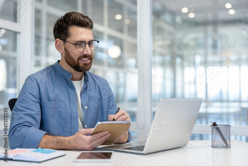 фотографія Positive bearded businessman working on a laptop, smiling and writing in a noteb