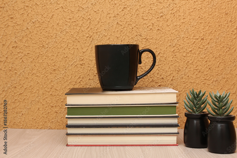 Fototapeta premium Black mug on top of a pile of books, on a wooden table, in front of orange color wall. Horizontal shot with copy space, perfect for study themes, course coffee break, and education contents.