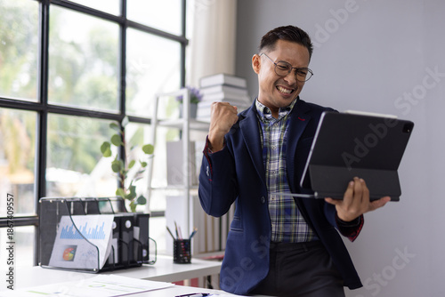 Young entrepreneur feeling euphoric while looking at tablet, celebrating successful business deal or good news in modern office.