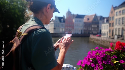 Woman tourist using smartphone on a bridge, browsing navigation app while exploring european canal city buildings in ghent, belgium, during a sunny travel day