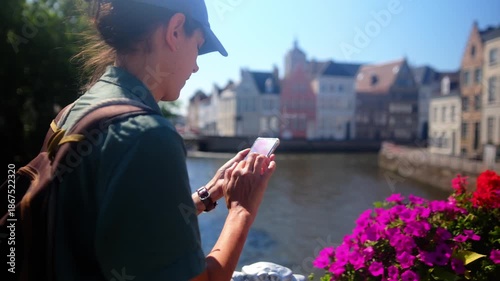 Woman tourist using smartphone on a bridge, browsing navigation app while exploring european canal city buildings in ghent, belgium, during a sunny travel day