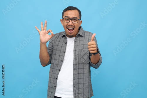 Handsome Asian man wearing casual clothes, showing okay sign hand gesture and thumbs up, smiling looking at camera, feeling satisfied, standing isolated on blue background.