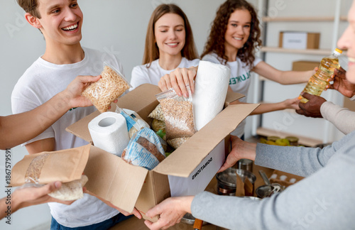 Young male and female volunteers happily pack food and personal hygiene products into boxes for donation. This charitable activity promotes community support and generosity among millennials.