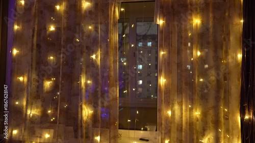 Night winter city view through window with warm glowing lights on curtains, apartment building outside, cozy home interior atmosphere and urban lifestyle concept