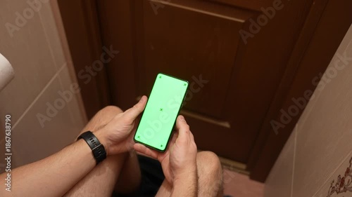 Top view of man using smartphone with green screen while sitting in restroom, hands scrolling on mobile phone, casual daily routine, technology mockup and modern lifestyle concept