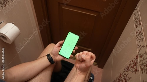 Top view of man using smartphone with green screen while sitting in restroom, hands scrolling on mobile phone, casual daily routine, technology mockup and modern lifestyle concept