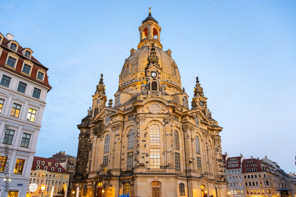 Obraz premium Dresden Frauenkirche features a large dome and ornate architecture. The church is surrounded by buildings and a clear sky during the evening. People visit the site during Christmas time.