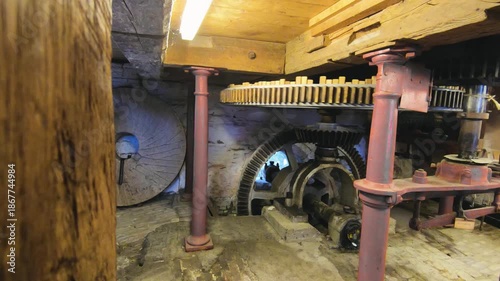 Interior of an old water mill showing a historic wooden and metal gear mechanism, cogwheels, shafts, and stone walls, industrial heritage machinery inside a rustic mill building