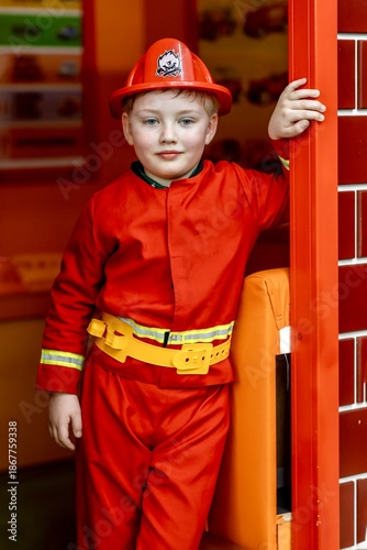Boy wearing a firefighter costume standing at the entrance of a play fire station in an indoor children’s entertainment center. Role play, childhood imagination, learning professions, safe indoor play