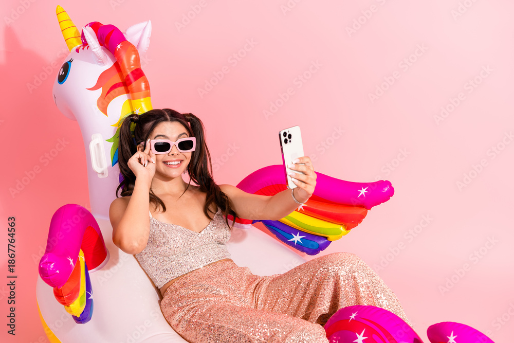 Obraz premium Young woman in glitter dress relaxing on a colorful unicorn floatie taking a selfie with a smartphone in a pink studio background