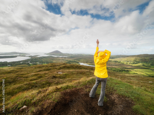 Tablou pe pânză Woman tourist in yellow jacket is standing on a hill with amazing nature scenery in the background with huge wild fields, ocean, mountain and cloudy sky