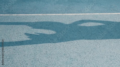 Cinematic shadows of basketball players on a bright blue textured outdoor court under harsh sunlight. A player holds a round ball high, ready for a shot. Minimalist, artistic composition with high con