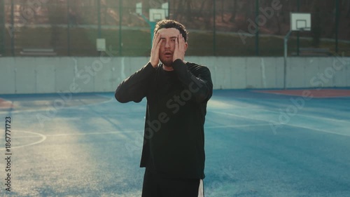 A young man in black sportswear stands on a blue basketball court with hands on his head. He looks shocked. Cinematic golden hour backlight creates a warm rim light. Moody urban sports atmosphere with