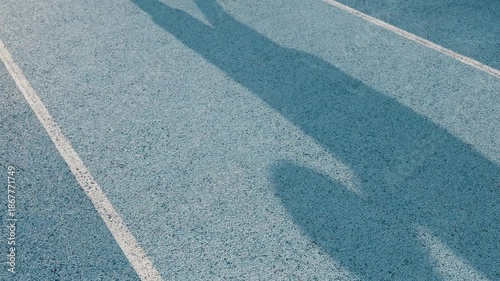 Slow motion striking dark shadows of an athlete holding a basketball cast onto a grainy blue outdoor court surface. Strong daylight creates high contrast against the sharp white line.