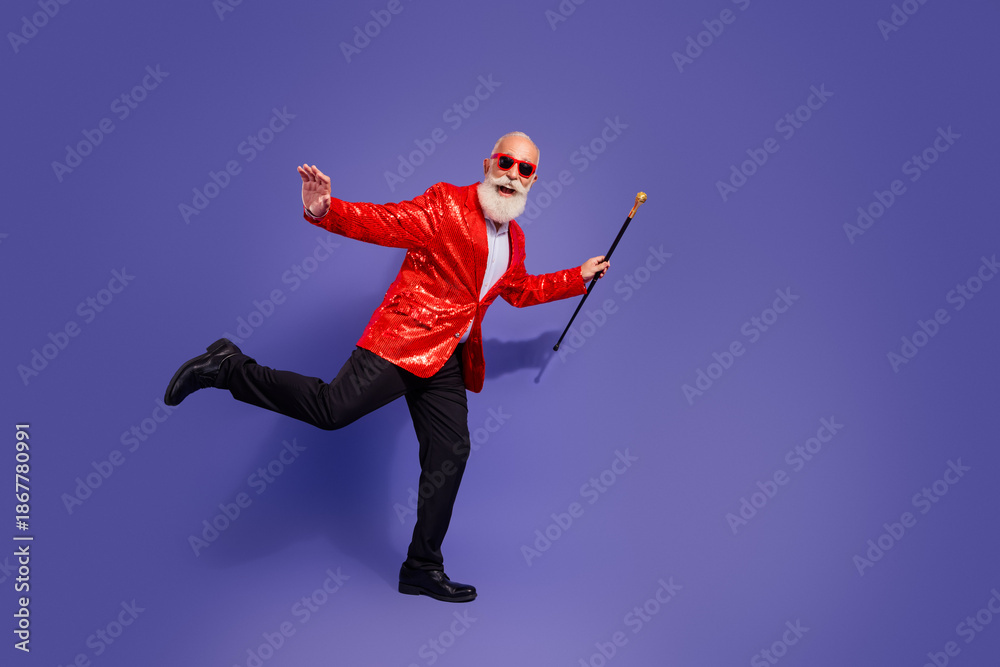 Fototapeta premium Energetic elderly showman in red sequin jacket dances with cane against purple background at party performance