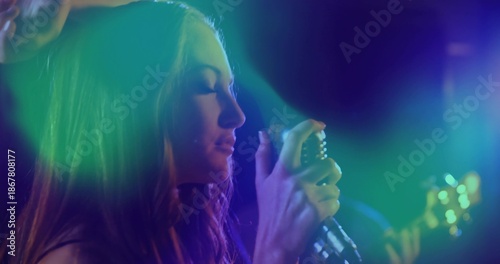 Singing female singer in dark outfit holding retro mic on stage with colored lights, blurred guitar