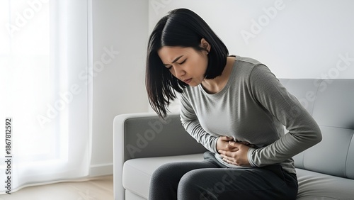 Young asian woman experiencing acute abdominal pain while sitting on a sofa at home