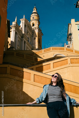 Obraz na płótnie Menton, France - March 25, 2025: Stylish woman posing on sunlit stairs with historic architecture and bell tower in a vibrant urban travel scene