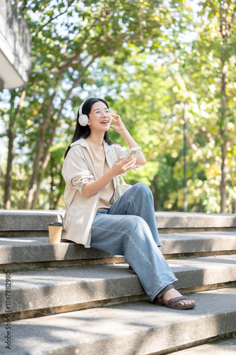 Free Download Glasses asian woman wearing headphones listening to music while sitting on concrete stairs in park. - GFXLoad