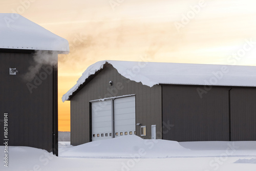 Minimal winter photo of metal pre-engineered industrial building with a snowbank on a roof after the snowfall. Yard with warehouses with steam from heater in sunset light, winter day..