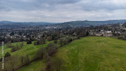 A wide view over open parkland in Bath, UK, with scattered trees, surrounding homes and the city spreading into rolling hills and countryside under a cloudy sky.