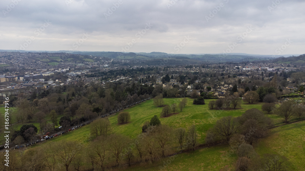 Obraz premium View across a park in Bath, UK, with bare trees, bordering roads and houses, and the city extending into rolling hills and countryside beneath an overcast sky.