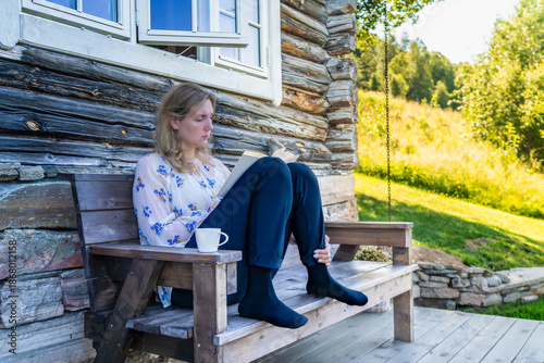 A woman sits on a wooden bench in front of a log cabin engrossed in a book. Enjoying her me time reading a book with a cup of tea in the Shade on a Sunny Day