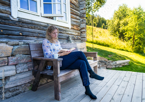 A beautiful woman sits on a wooden bench in front of a log cabin engrossed in a book. Enjoying her me time reading a book with a cup of tea in the Shade on a Sunny Day