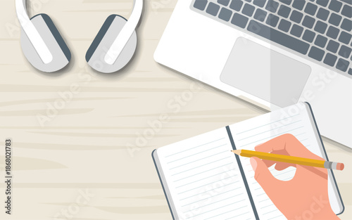 A hand holding a pencil writes in a notebook on a desk with a laptop and headphones nearby